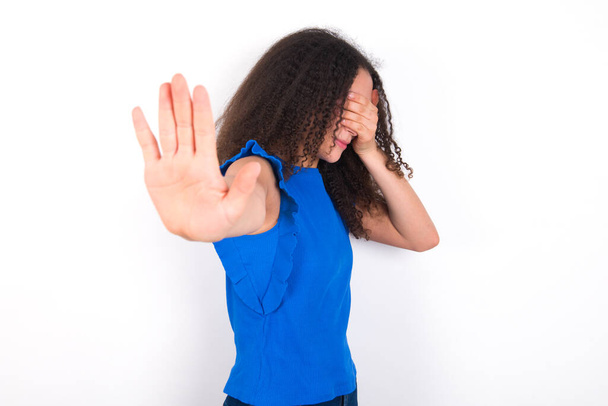 Nastolatka z afro fryzury noszenie niebieskiej koszulki na białym tle zakrywa oczy dłonią i robi gest stop, stara się ukryć przed wszystkimi. - Zdjęcie, obraz
