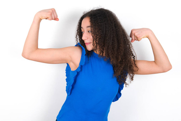 Teenager-Mädchen mit Afro-Frisur trägt blaues T-Shirt über weißem Hintergrund und zeigt stolz lächelnde Armmuskeln. Fitnesskonzept. - Foto, Bild