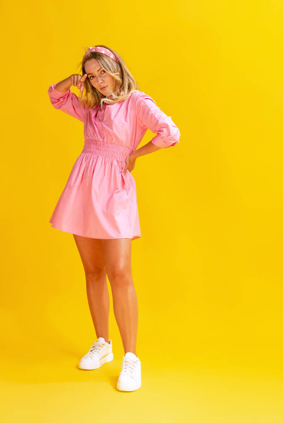 Portrait of beautiful young woman in cute pink dress posing isolated over yellow studio background. Concept of beauty, emotions, facial expression, lifestyle, fashion, youth culture - 写真・画像