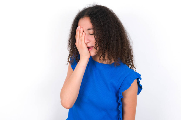 Menina adolescente com penteado afro vestindo camiseta azul sobre fundo branco bocejo cansado cobrindo metade do rosto, olho e boca com a mão. Cara dói na dor. - Foto, Imagem