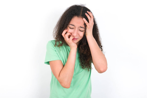 Doleful desperate crying Teenager girl with afro hairstyle wearing white T-shirt over green background , looks stressfully, frowns face, feels lonely and anxious - Photo, image