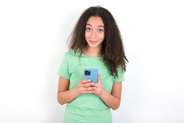 Дівчина-підліток з зачіскою на афро в білій футболці над зеленим тлом насолоджується віддаленим спілкуванням, використовує мобільний телефон, панує швидкий безмежний Інтернет, приємно посміхається, робить покупки онлайн
, - Фото, зображення