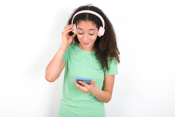 Glückliche Teenager-Mädchen mit Afro-Frisur tragen weißes T-Shirt über grünem Hintergrund fühlt sich gut, während konzentriert auf dem Bildschirm des Smartphones. Menschen, Technologie, Lebensstil - Foto, Bild