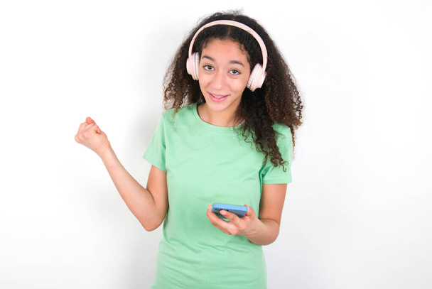Positive Teenager girl with afro hairstyle wearing white T-shirt over green background holds modern cell phone connected to headphones, clenches fist from good emotions, exclaims with joy, - Photo, Image