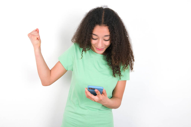 Teenager girl with afro hairstyle wearing white T-shirt over green background holding in hands cell and rising his fist up being excited after reading good news. - Foto, imagen