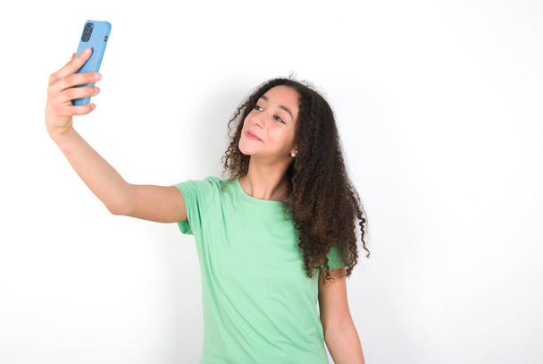 Teenager-Mädchen mit Afro-Frisur trägt weißes T-Shirt über grünem Hintergrund, lächelt und macht ein Selfie, um es in ihren sozialen Medien zu posten. - Foto, Bild