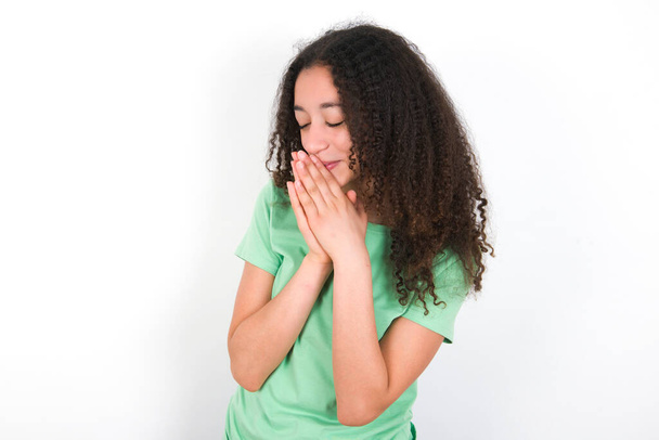 Overjoyed Teenager girl with afro hairstyle wearing white T-shirt over green background laughs joyfully and keeps palms pressed together hears something funny - 写真・画像
