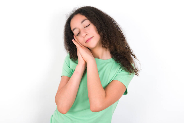 Teenager girl with afro hairstyle wearing white T-shirt over green background leans on pressed palms closes eyes and has pleasant smile dreams about something - Valokuva, kuva