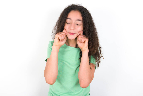 Pleased Teenager girl with afro hairstyle wearing white T-shirt over green background with closed eyes keeps hands near cheeks and smiles tenderly imagines something very pleasant - Фото, изображение