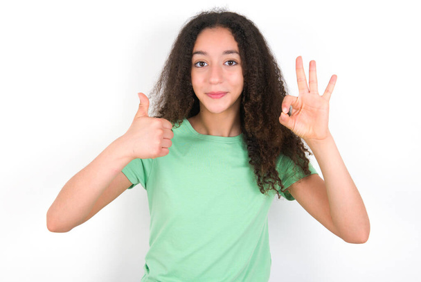 Teenagermädchen mit Afro-Frisur trägt weißes T-Shirt über grünem Hintergrund und sieht glücklich, unbeschwert und positiv aus, gestikuliert Sieg oder Frieden mit einer Hand - Foto, Bild