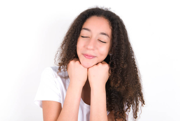 Cheerful Teenager girl with afro hairstyle wearing white T-shirt over white background has shy satisfied expression, smiles broadly, shows white teeth, People emotions - 写真・画像