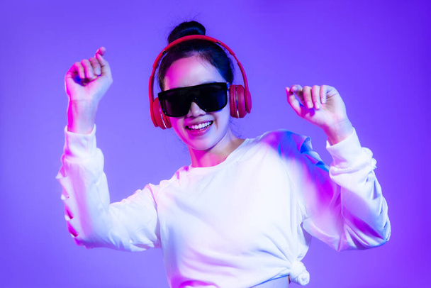 Νεαρή ασιάτισσα με λευκό φούτερ μόδας φορώντας γυαλιά ηλίου και κόκκινα ακουστικά ακούει μουσική και χορεύει στο μωβ φόντο. - Φωτογραφία, εικόνα