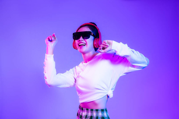 asian woman in white fashion crop sweatshirt wearing sunglasses and red headphones listen to music and dancing isolated purple background. - Foto, afbeelding