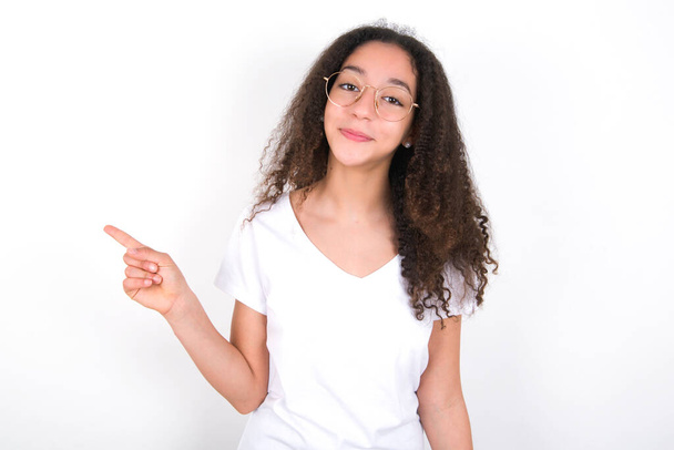 Positives Teenager-Mädchen mit Afro-Frisur trägt weißes T-Shirt über weißem Hintergrund mit zufriedenem Gesichtsausdruck zeigt oben rechts gutes Angebot schlägt vor, auf Link zu klicken - Foto, Bild