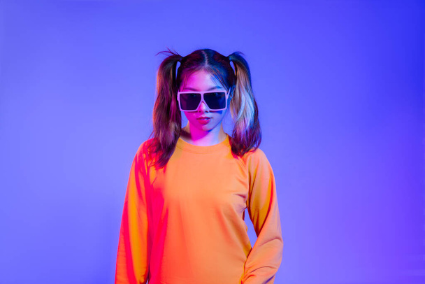 Young asian girl in yellow sweatshirt wearing sunglasses posing on blue screen background. - Foto, Imagen