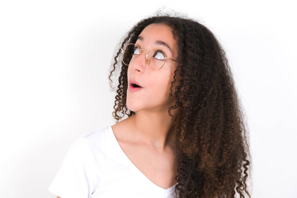 Geschokt tiener meisje met afro kapsel dragen witte T-shirt over witte achtergrond kijken lege ruimte met open mond schreeuwen: Oh mijn God! Ik kan dit niet geloven.. - Foto, afbeelding
