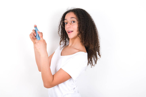 Nice addicted cheerful Teenager girl with afro hairstyle wearing white T-shirt over white background using gadget playing network game - Photo, image