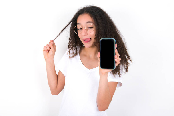 Photo of nice pretty Teenager girl with afro hairstyle wearing white T-shirt over white background demonstrate phone screen hold hair tails - Foto, Imagem