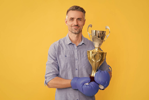usmívající se dospělý muž v boxerských rukavicích drží zlatý pohár na žlutém pozadí, vítězství. - Fotografie, Obrázek