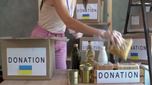 Donation of food for refugees from Ukraine affected by the war, support for the victims, helping people, charity, a box with the Ukrainian flag. a female volunteer puts groceries in a box. - Filmati, video
