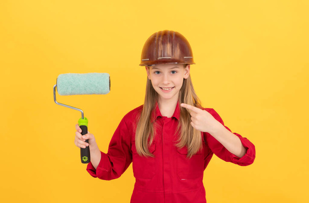 happy teen child in builder hard hat with paint roller on yellow wall. point finger. - Fotoğraf, Görsel