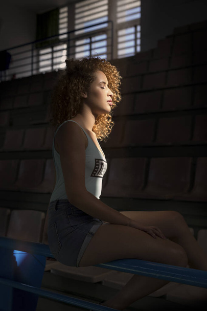 Side view of tranquil female with curly hair in denim shorts sitting under sunlight with closed eyes in dark stadium - Photo, Image