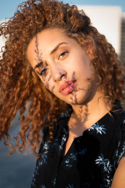 Beautiful young female with curly red hair looking at camera while standing under sunlight with shadow of twigs on face - Foto, Imagem