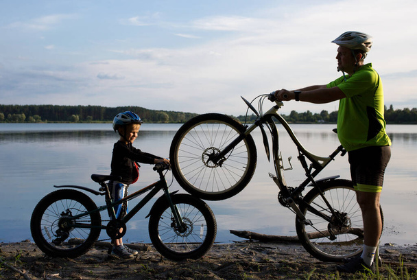 Silhouettes of an adult man and a boy with two bicycles in helmets near the lake at sunset. cycling, healthy lifestyle. Be like dad. Family pastime, dad and son ride a bike - Photo, Image