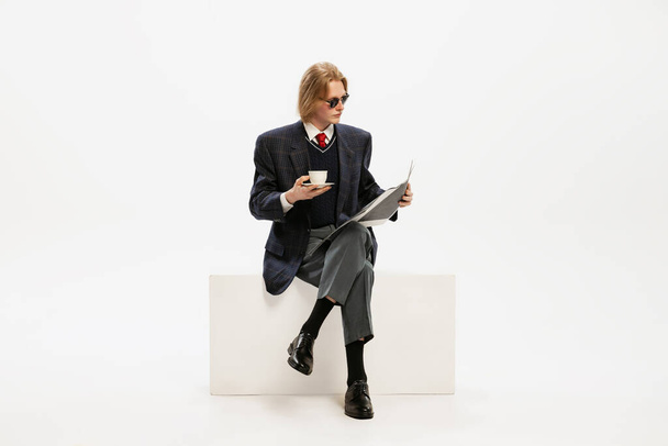 Πορτρέτο του κομψού νεαρού άνδρα σε σύνολο μαύρο στολή πίνοντας καφέ, διαβάζοντας εφημερίδα, θέτοντας απομονωμένη σε λευκό φόντο στούντιο. Έννοια της ρετρό μόδας, στυλ, νεανική κουλτούρα, συναισθήματα, ομορφιά, διαφήμιση - Φωτογραφία, εικόνα