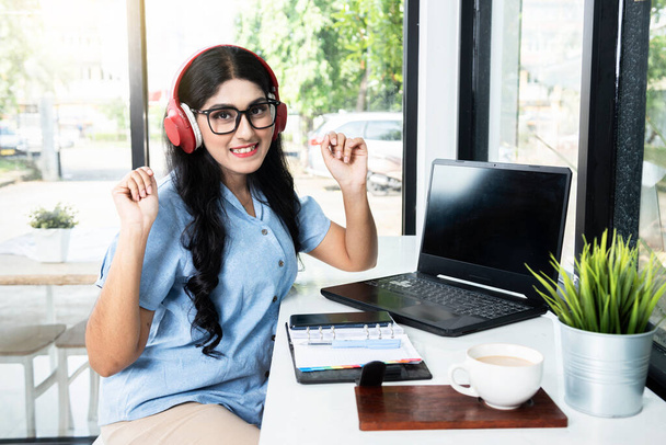 Asian woman with eyeglass listening to music with headphones with a laptop and a notebook with a mobile phone and a cup of coffee on the table in the coffee shop - Photo, Image
