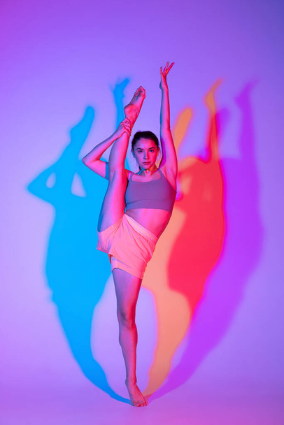 Flexibility. Young sportive girl standing in twine isolated on gradient studio background in neon light. athletic and graceful. Modern sport, action, fitness, yoga, motion, youth concept. - Zdjęcie, obraz