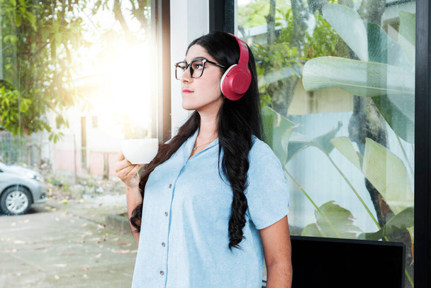 Asian woman with eyeglass listening to music with headphones and holding a cup of coffee with a laptop on the table in the coffee shop - Foto, Imagem