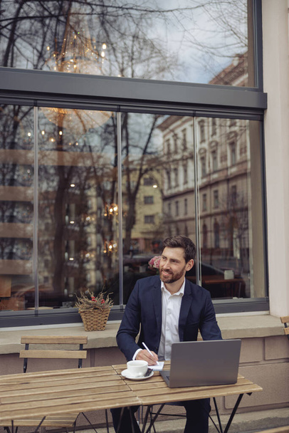 Ελκυστικό κομψό άνθρωπος κρατώντας σημειώσεις, ενώ εργάζονται σε φορητό υπολογιστή στο καφέ έξω. Συγγραφέας σε διάλειμμα για καφέ. - Φωτογραφία, εικόνα