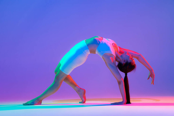 Stretching. Studio shot of young flexible girl in fitness sport uniform practicing isolated on gradient pink-purple background in neon. Modern sport, action, fitness, yoga, motion, youth concept. - Фото, зображення