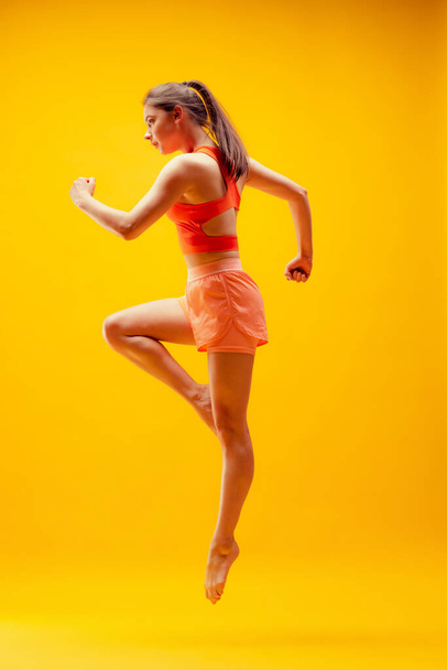 Running, jumping. Full-length portrait of young sportive girl in motion isolated on bright yellow background. Modern sport, action, motion, summer, vacation, youth concept. - Foto, Bild