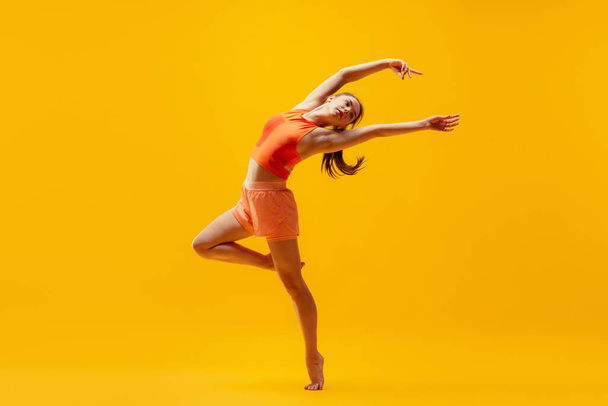 Portrait of young slim, flexible girl in shorts doing stretching exercises isolated on bright yellow background. Modern sport, action, motion, summer, vacation, youth concept. - Foto, afbeelding