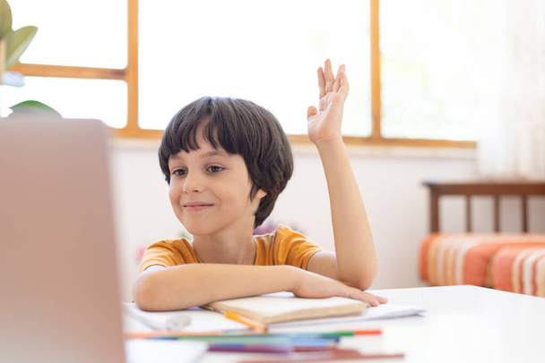 мальчик поднимает руку во время урока онлайн дистанционного обучения. мальчик с компьютером общался через видеоконференцию с учителем и классной группой. Ребенок общается онлайн. - Фото, изображение