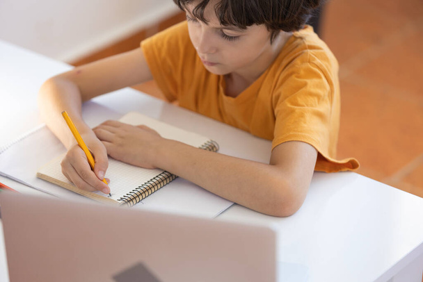 een jongen schrijft en tekent in een notitieboekje tijdens een online les. Online leren op afstand. een jongen met een computer communiceert via videoconferentie met een leraar en een klasgroep.  - Foto, afbeelding