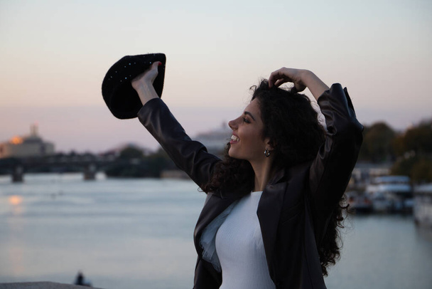 Young, beautiful woman with dark, curly hair opening her arms in the air. In the background part of the city and the river guadalquivir in the golden hour. Tourism and holiday concept. - Zdjęcie, obraz