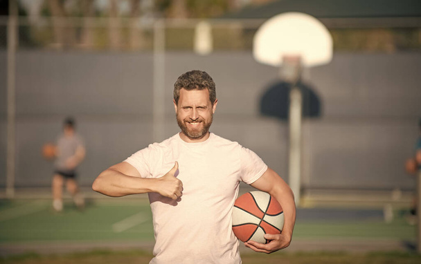 wesoły muskularny człowiek sportowiec koszykarz trzymać piłkę na świeżym powietrzu pokazując gest kciuk w górę, sukces sportowy. - Zdjęcie, obraz
