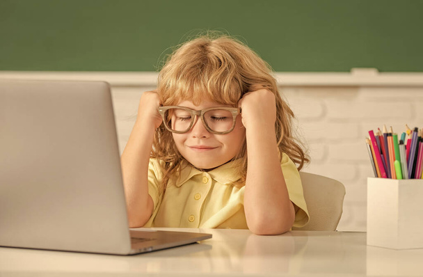 сонний хлопчик в окулярах навчається онлайн в шкільному класі з ноутбуком, освіта
. - Фото, зображення