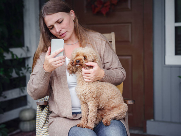 sonriente mujer caucásica de mediana edad en chaqueta marrón toma selfie o se comunica a través de video por amigos con su caniche mascota en la terraza del patio trasero. divertido, acogedor y cómodo estilo de vida otoñal - Foto, imagen
