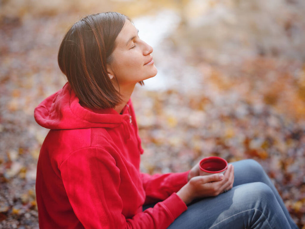 Travel and road trip concept at autumn. Adventure and active lifestyle in nature. Asian woman in red hoodie drinking tea from thermos sitting by river in forest. Wanderlust concept. - Photo, Image