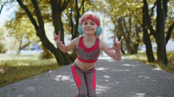 Sportos fitness sport futó gyermek lány edzés gyakorlat zenét hallgatni a fejhallgató tánc kamera szórakozás. Edző kardió a parkban. Kisgyermekek gyerek kocogó élvezi vicces divat tánc. - Fotó, kép
