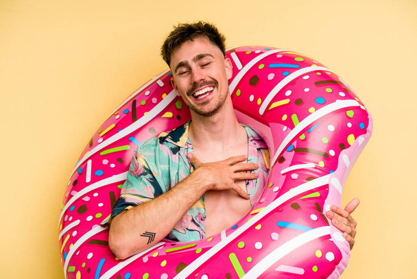 Young caucasian man holding an inflatable donut isolated on yellow background laughs out loudly keeping hand on chest. - Foto, immagini