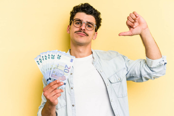 Young caucasian man holding banknotes isolated on yellow background feels proud and self confident, example to follow. - Zdjęcie, obraz