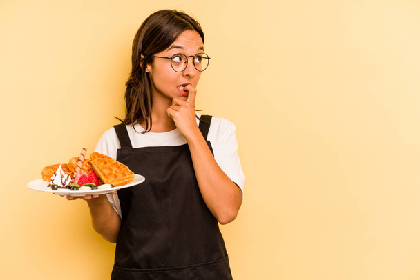 Young hispanic dependent woman holding waffles isolated on yellow background relaxed thinking about something looking at a copy space. - Photo, Image