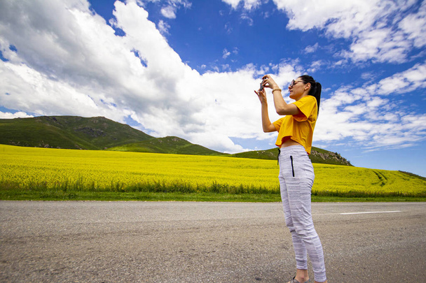 girl on the road, in a yellow T-shirt, taking a photo in a flowering field, against the backdrop of mountains - Fotoğraf, Görsel