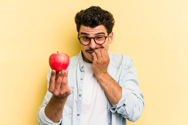 Молодой кавказский мужчина держит яблоко на желтом фоне, кусает ногти, нервничает и очень беспокоится. - Фото, изображение