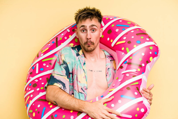 Young caucasian man holding an inflatable donut isolated on yellow background shrugs shoulders and open eyes confused. - Foto, Bild
